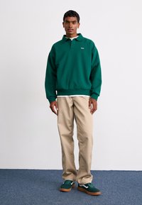 Pullover verde com colarinho e logotipo bordado, combinado com calças bege e ténis verdes com detalhes brancos. Textura suave por todo o lado.