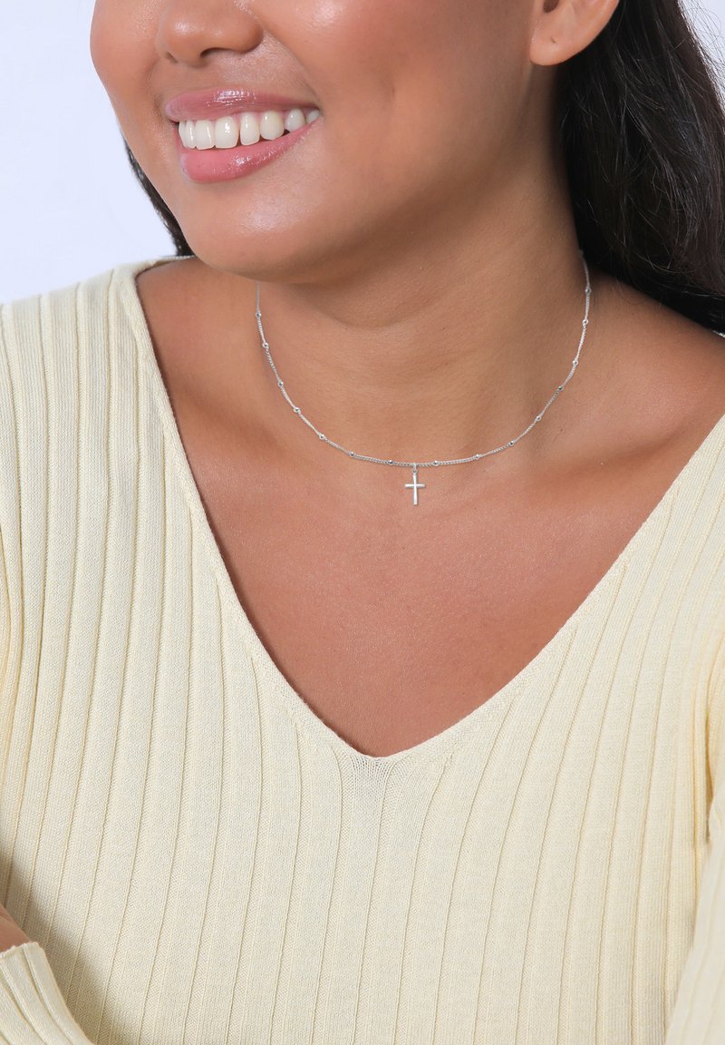 Silver necklace with small beaded accents and a central cross pendant, worn with a light yellow ribbed top.