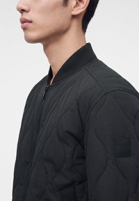 Vue de profil d'une personne portant une veste matelassée noire avec un col côtelé et un patch logo BOSS sur la manche.