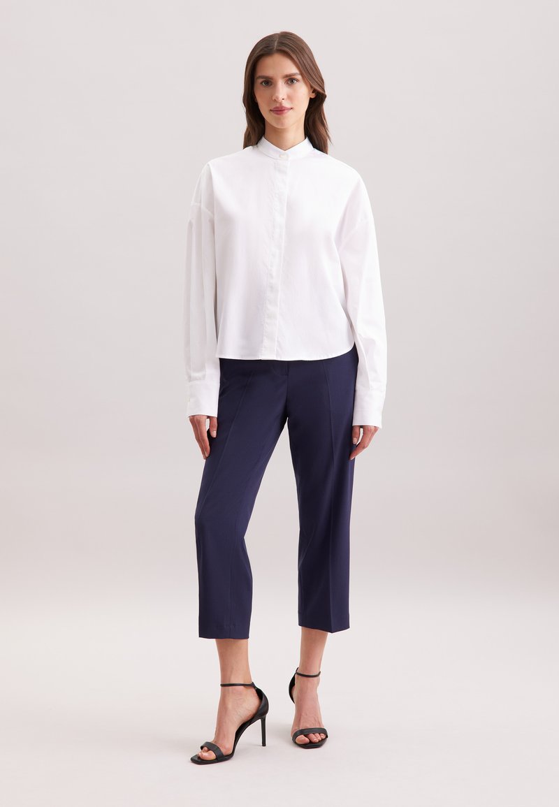 Camicia bianca a maniche lunghe con colletto alla cinese, abbinata a pantaloni blu scuro a 3/4 e sandali neri con tacco. Tessuto liscio, vestibilità su misura.