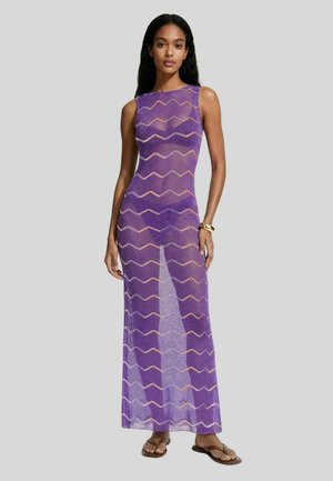 Donna che indossa un vestito viola trasparente senza maniche, lungo fino al pavimento, con motivi a zigzag, abbinato a sandali e un braccialetto d'oro al polso destro.