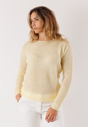Donna con capelli mossi, che indossa un maglione di maglia giallo chiaro e pantaloni bianchi, in piedi con una mano in tasca, mentre guarda di lato.