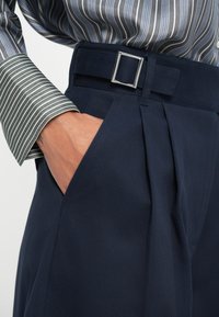 Pantalons navy ajustés avec un devant plissé, dotés d'une taille plate et d'un détail en boucle argentée. Manches de chemise rayées en vert et blanc.
