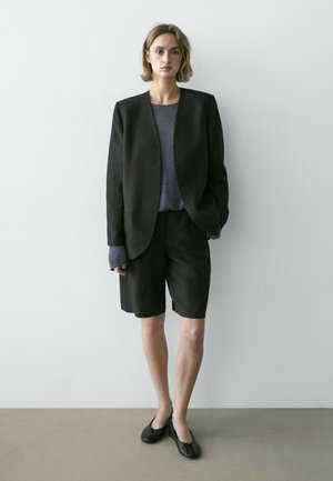 Donna in piedi contro il muro che indossa un blazer nero oversize, pantaloncini neri al ginocchio, maglione grigio a maglia e ballerine nere.