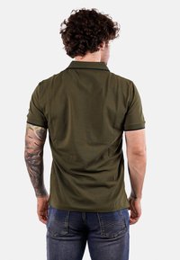 Camiseta polo verde oliva hecha de una tela suave, con cuello, mangas cortas y ribetes de contraste. Combinada con unos vaqueros azules.