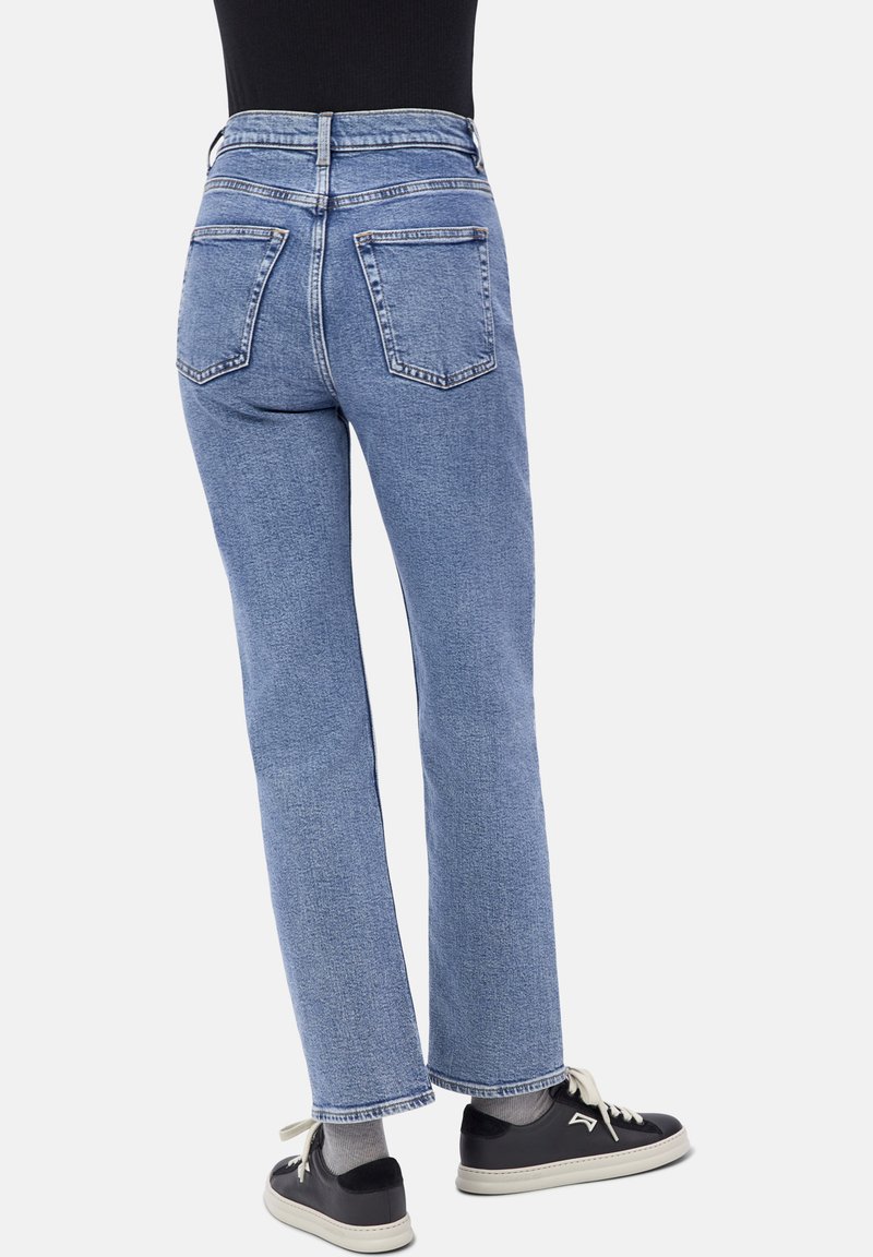 Personne portant un jean droit bleu clair, un haut noir, des chaussettes grises et des baskets noires avec des semelles et des lacets blancs, se tenant sur un fond blanc.