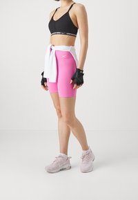 Brassière de sport noire, short rose, haut blanc noué à la taille. Gants sans doigts noirs, baskets rose clair. Le tissu est extensible et lisse.