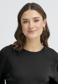 Sweat-shirt noir à col rond et tissu texturé. Le modèle a les cheveux bruns mi-longs et porte des boucles d'oreilles créoles dorées.