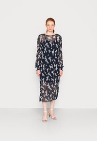 Vestido azul-marinho transparente com padrão floral branco, mangas bufantes longas e um corte solto, que atinge a metade da panturrilha, combinado com sandálias de tiras claras.