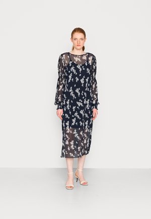 Vestido azul-marinho transparente com padrão floral branco, mangas bufantes longas e um corte solto, que atinge a metade da panturrilha, combinado com sandálias de tiras claras.