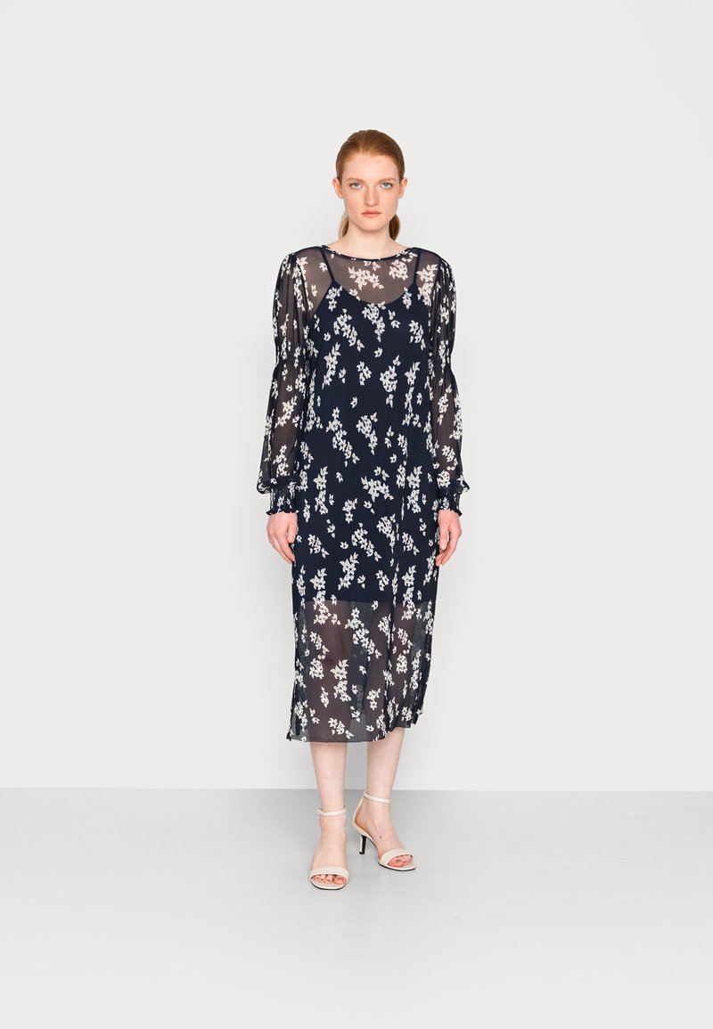Vestido azul-marinho transparente com padrão floral branco, mangas bufantes longas e um corte solto, que atinge a metade da panturrilha, combinado com sandálias de tiras claras.