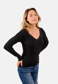 Donna con lunghi capelli biondi, che indossa un maglione nero a scollo a V con bordi in pizzo e jeans blu, in piedi con le mani sui fianchi su uno sfondo bianco.