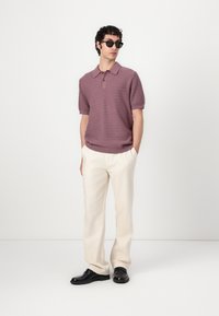 Short-sleeve, textured purple polo with a collar, paired with cream trousers and black shoes, set against a neutral background.