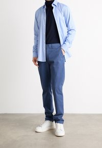 Camicia button-up azzurro chiaro sopra un dolcevita nero, abbinata a pantaloni blu scuro e sneakers bianche. Tessuto liscio e design aderente.