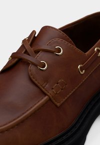 Καφέ δερμάτινο μοκασίνι με χρυσούς κρίκους και κορδόνια από το ίδιο δέρμα, με λεπτομερείς ραφές και μαύρη σόλα.