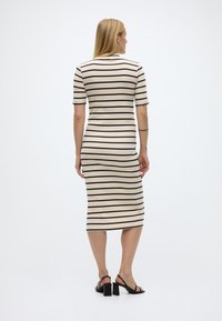 Γυναίκ α με μακριά ξανθά μαλλιά, ντυμένη με μπεζ μεσαίο φόρεμα με μαύρες οριζόντιες ρίγες και μαύρα σανδάλια με χοντρό τακούνι, γυρισμένη προς τα πίσω.