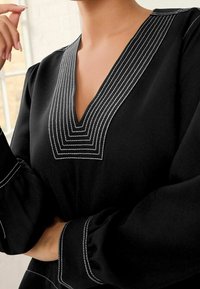 Schwarze Bluse aus Stoff mit V-Ausschnitt, der weiße geometrische Stickereien am Ausschnitt und puffige Ärmel mit passenden Details aufweist.