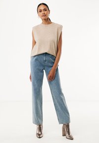 Top sin mangas beige, con cuello alto, combinado con jeans rectos de color azul claro que presentan una textura moteada y botas de tobillo metálicas con tacón.