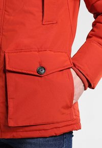 Červená prošívaná bunda s velkou kapsou vpředu zavíranou na knoflík, texturovanou látkou a žebrovanými manžetami na koncích rukávů.