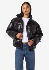 Juoda pūkinė striukė su blizgiu paviršiumi, turinti aukštą apykaklę, užtrauktuką ir laisvo dydžio kirpimą. Derinama su šviesiai mėlynomis džinsomis.