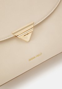 Sac à main en cuir beige avec fermoir triangulaire doré et petit logo "ANNA FIELD" doré embossé sous le rabat.