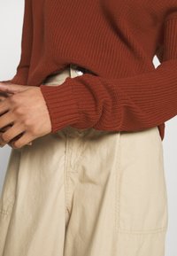 Rust-colored knitted sweater with ribbed texture, featuring long sleeves and a relaxed fit, paired with light beige high-waisted pants.