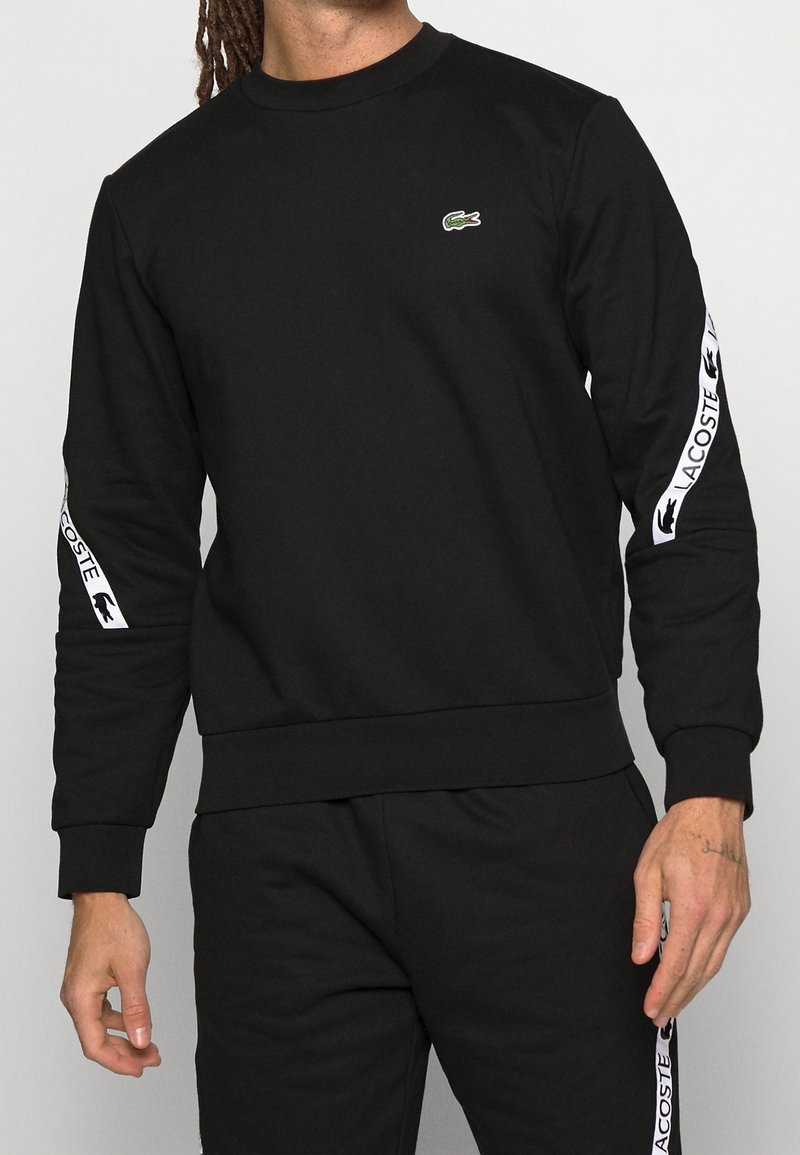 Homme portant un sweat-shirt noir Lacoste et un pantalon assorti avec des bandes blanches du logo Lacoste sur les manches et les jambes.