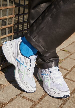 Białe sportowe trampki z niebieskimi i zielonymi akcentami, noszone z jasnoniebieskimi skarpetkami i czarnymi skórzanymi spodniami, stojące na wybrukowanym podłożu obok metalowej kratki.