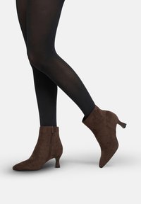 Botines de ante marrón con punta afilada, cierre con cremallera y tacón modesto, mostrados junto a medias negras.