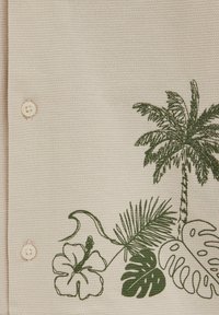 Tissu beige texturé avec deux boutons beiges et un imprimé tropical vert représentant un palmier, une fleur d’hibiscus et diverses feuilles.