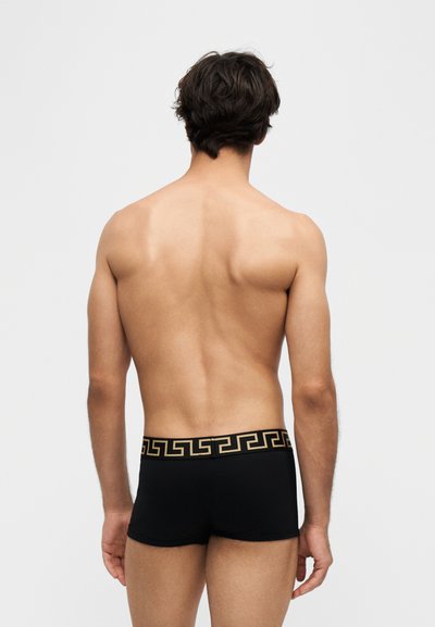 Homme aux cheveux courts et foncés vu de dos, portant un boxer noir avec une ceinture à motif clé grecque dorée sur fond blanc.