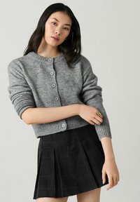 Gray knitted cardigan with ribbed hem, featuring five buttons at the front. Paired with a black pleated skirt, creating a layered look.