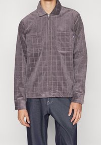 Chaqueta de pana en un tono púrpura apagado, con un patrón de cuadrícula, cierre frontal con cremallera y dos bolsillos en el pecho. Textura suave y corte relajado.