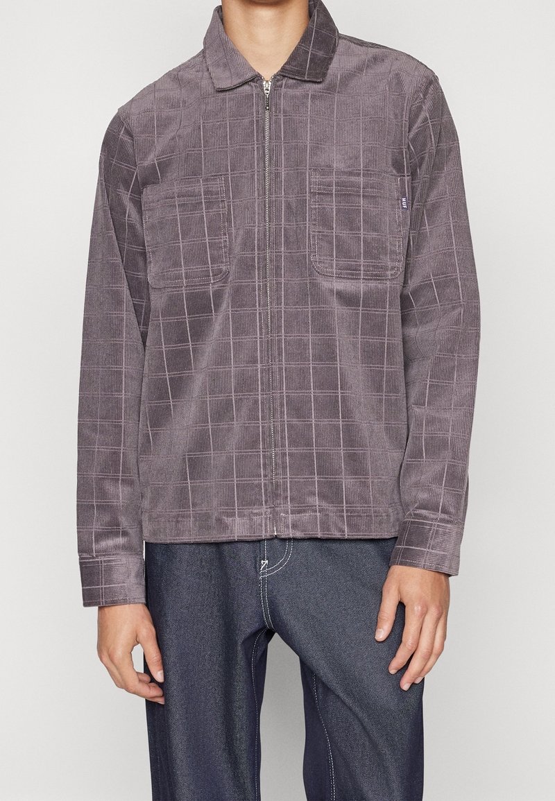 Chaqueta de pana en un tono púrpura apagado, con un patrón de cuadrícula, cierre frontal con cremallera y dos bolsillos en el pecho. Textura suave y corte relajado.