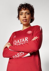 Långärmad fotbollströja i röd med vita detaljer. Har logotyper: "QATAR AIRWAYS" och "PARIS." Slät tyg, atletisk passform, rund halsringning.