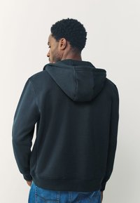 Čierna mikina s kapucňou na šnúrku, vyrobená z mäkkého materiálu. Má rebrované manžety a lem, s voľným strihom. Pohľad zozadu ukazuje hladkú textúru.