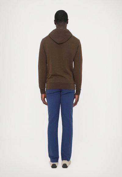 Középméretű barna kapucnis pulóver zsinóros kapucnival. Kék melegítőnadrággal és világos cipővel párosítva, hátulnézetben.