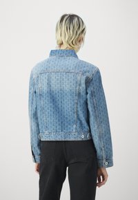 Persona con cabello rubio corto que lleva una chaqueta vaquera azul con estampado y pantalones negros, de pie de espaldas sobre un fondo liso.