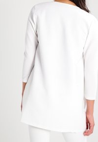 Bílý svetr s dlouhým rukávem, vyrobený z texturované látky, s kulatým výstřihem a bočními rozparky. Zadní část je mírně delší než přední.