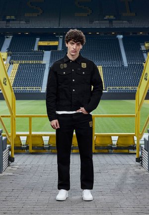 Młody mężczyzna w czarnej kurtce i spodniach z żółtymi sportowymi logo stoi na schodach stadionu zwrócony do kamery, z zielonym boiskiem piłkarskim w tle.