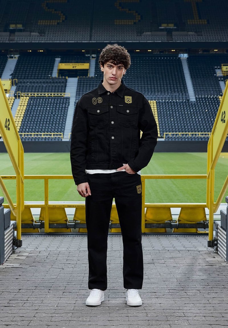 Jeune homme en veste et pantalon noirs avec des logos sportifs jaunes, debout sur les marches du stade face à la caméra, avec un terrain de football vert derrière.