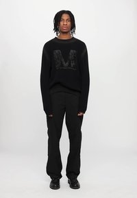 Svart stickad tröja med ribbad textur som har en stor stiliserad bokstavsdesign, ihopparat med svarta byxor och svarta stövlar.