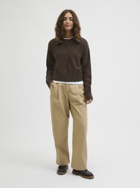 Jovem mulher com cabelo castanho ondulado, vestindo um cardigan castanho com botões, uma camiseta branca, calças beiges de perna larga e sapatos castanho-escuros, de pé.