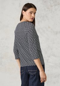 Topp med trekvartslång ärm i blått och vitt geometriskt diamantmönster, tillverkad av lättviktig tyg, ihop med mörka jeans.