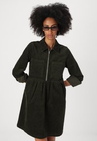 Rochie din catifea de culoare verde închis, cu fermoar în față, guler și două buzunare pe piept. Formă în A, cu talie strânsă și mâneci răsucite.