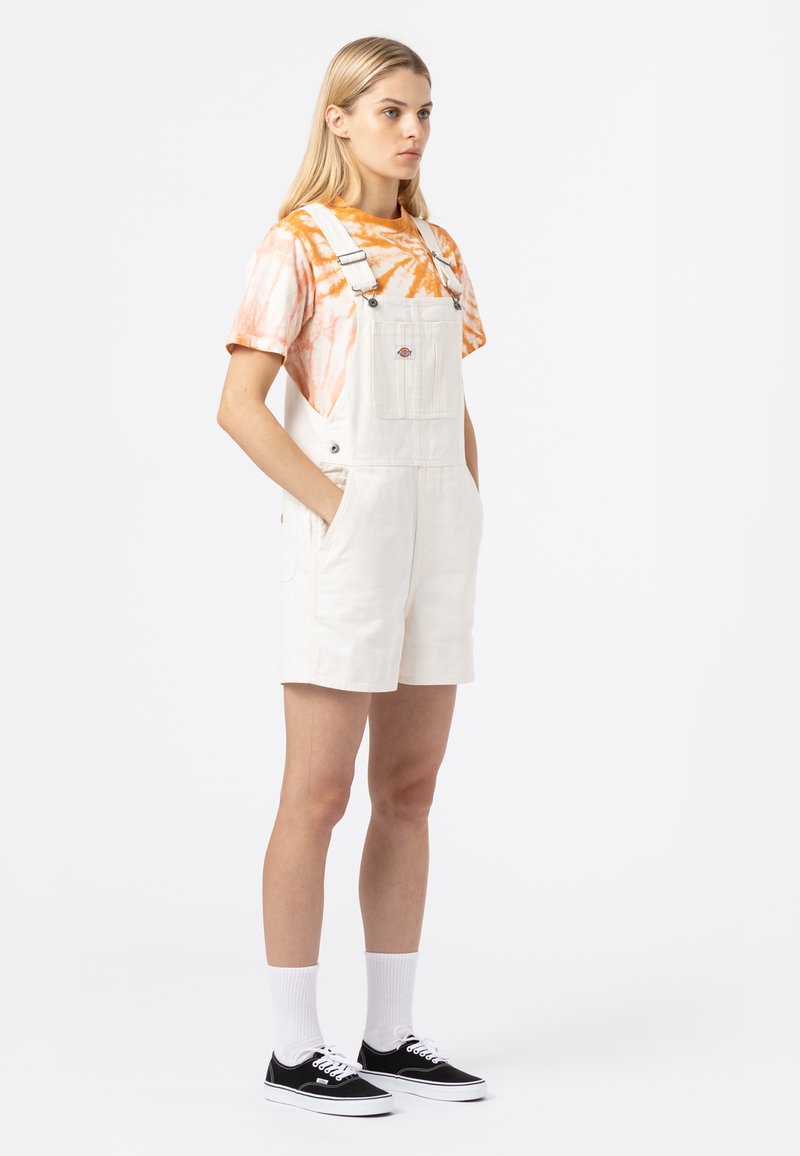 Hvite denim overall shorts med justerbare stropper, sidelommer og et logopatch. Kombinert med en kortermet oransje tie-dye skjorte og svarte sko.