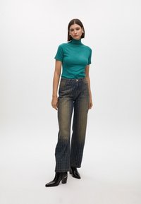 Teal short-sleeve turtleneck top paired with dark denim jeans featuring gold dot embellishments. Model wears black heeled boots.