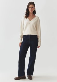 Pulover tricotat de culoare filă cu decolteu în V și design cu tricotaj tip cablu, asortat cu blugi drepti de culoare albastru închis și cizme scurte maro. Look simplu și casual.