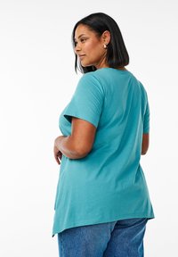 Teal kortærmet T-shirt med scoop-udskæring, der har en rund kant og en afslappet pasform, båret med højtaljede denimjeans.
