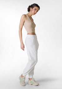 Beige geribbelde tanktop, witte joggingsbroek met koord, en lichte schoenen met groene accenten. Model staat in profielweergave, gladde textuur.